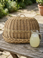 Handwoven wicker bread cloche with tray on a weathered garden table beside a glass jug of lemonade on a sunny patio