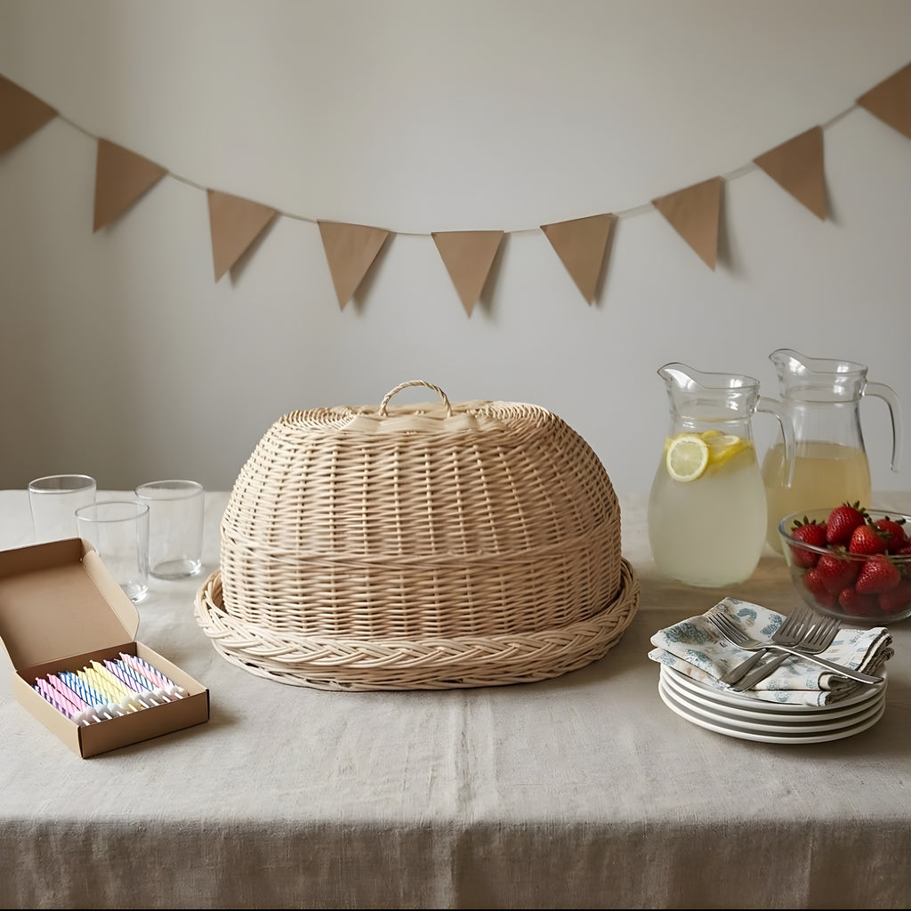 Oval wicker bread cloche with tray on a party table with lemonade jugs, strawberries, napkins and a box of birthday candles