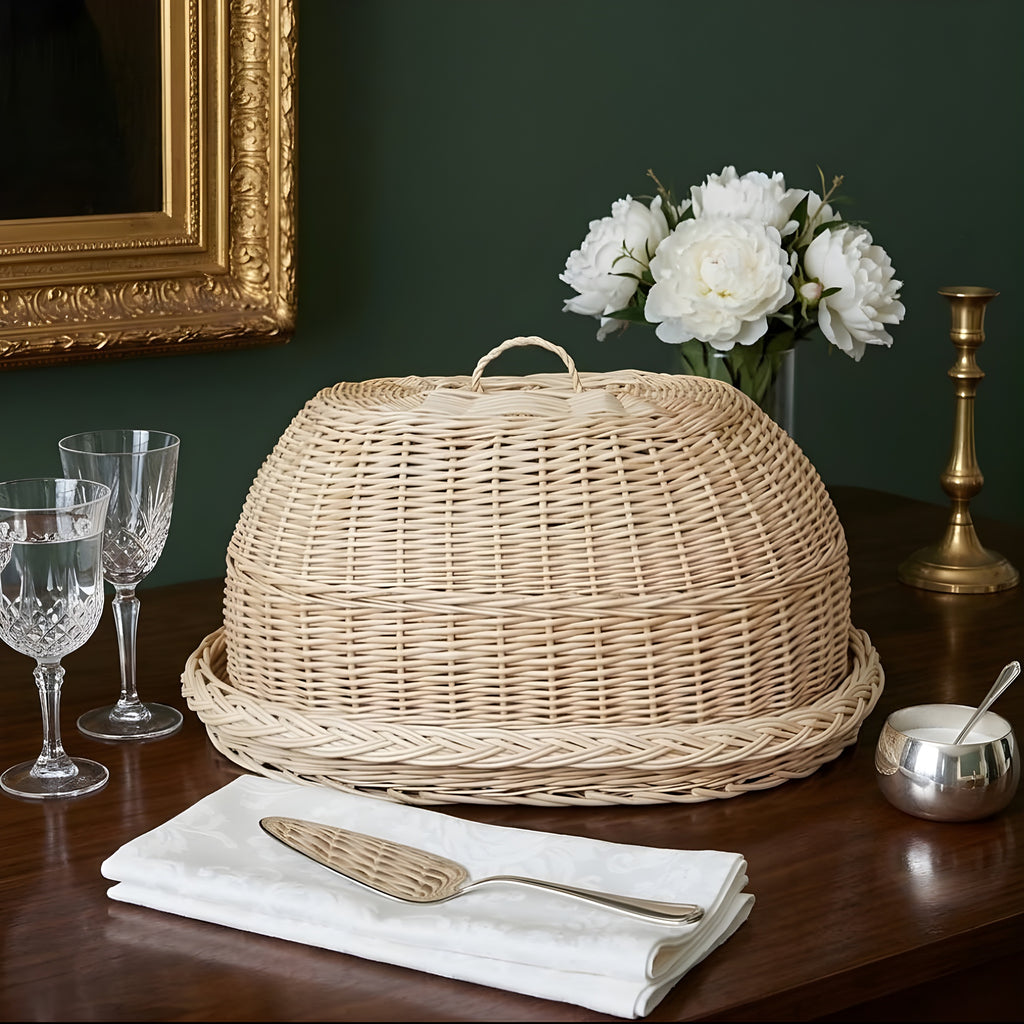 Handwoven willow bread cloche with a loop handle on its matching oval tray on a dark wooden table with peonies and crystal glasses