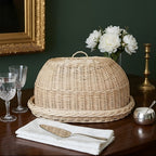 Handwoven willow bread cloche with a loop handle on its matching oval tray on a dark wooden table with peonies and crystal glasses