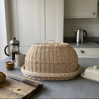 Wicker bread cloche with loop handle on its oval tray on a grey kitchen counter beside a coffee press, butter dish and bread knife