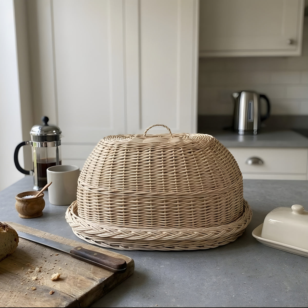 Wicker bread cloche with loop handle on its oval tray on a grey kitchen counter beside a coffee press, butter dish and bread knife