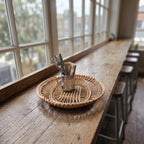 Handwoven willow openwork tray holding a metal cup of cutlery on a long wooden café counter by tall windows.