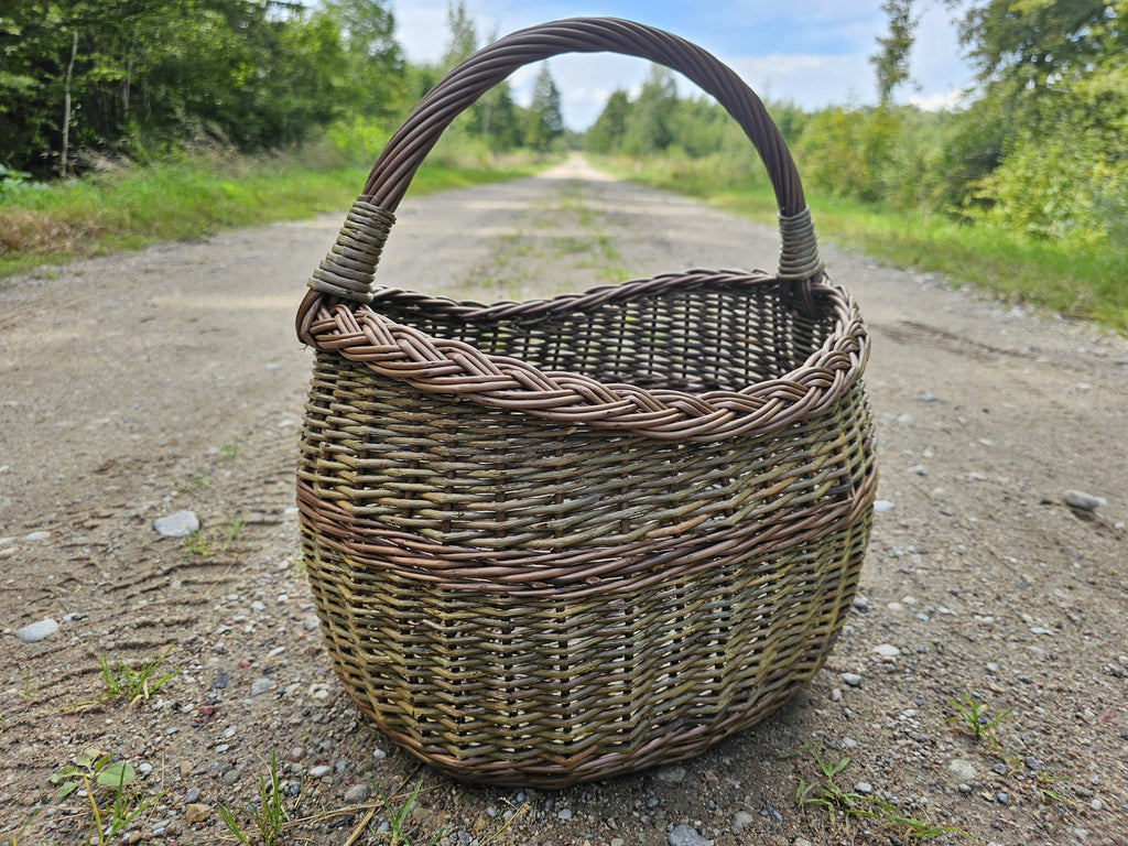 Handwoven willow market basket photographed head-on on a gravel forest track, the arched handle rising above the rounded body.