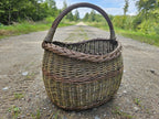 Handwoven willow market basket photographed head-on on a gravel forest track, the arched handle rising above the rounded body.