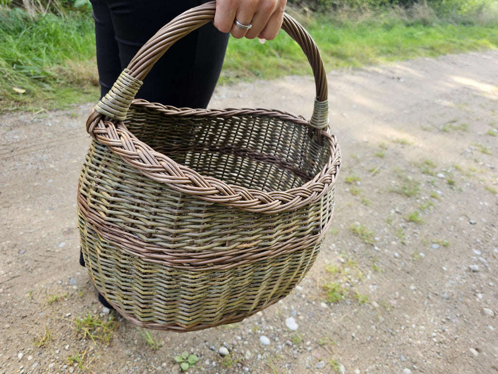 The wicker basket carried in one hand above a gravel path, showing its size against a person and how the handle sits in the grip.