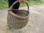 The wicker basket carried in one hand above a gravel path, showing its size against a person and how the handle sits in the grip.