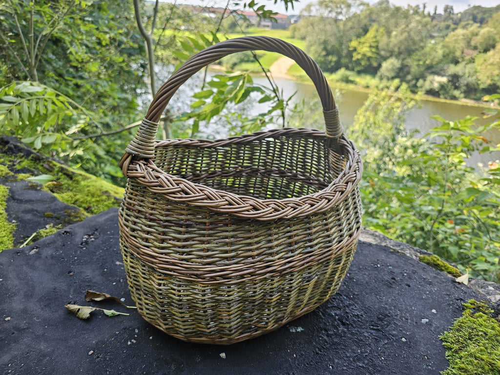 Three-quarter view of the willow basket on riverside stone, the river visible beyond the moss and rock.