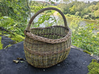 Three-quarter view of the willow basket on riverside stone, the river visible beyond the moss and rock.