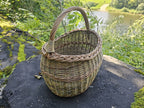 Handwoven willow basket standing on dark stone with the river behind, showing the full height including the handle.