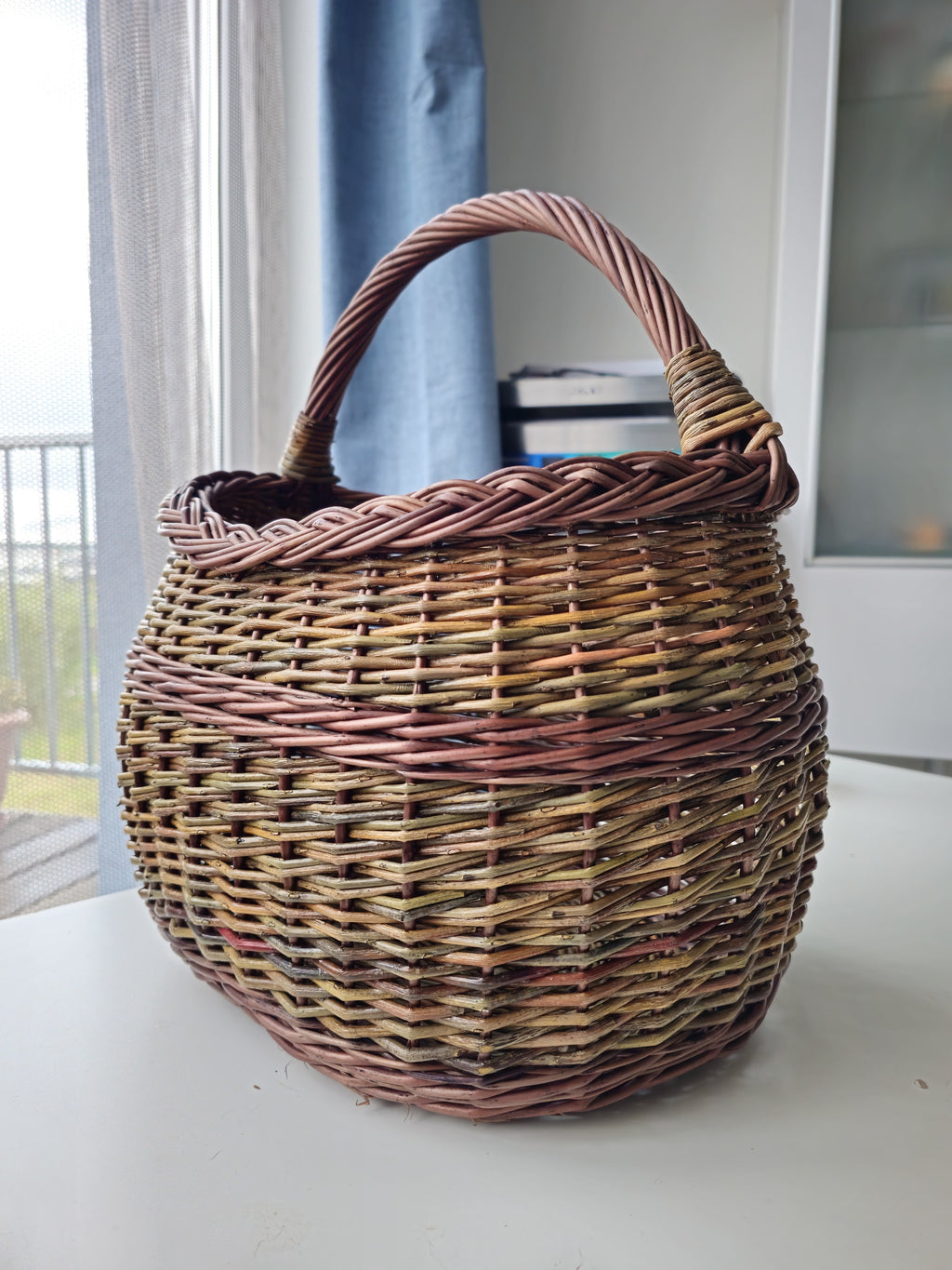 Handwoven unpeeled willow market basket indoors on a white table by a window, the rim and handle seen against the light.