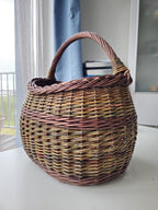 Handwoven unpeeled willow market basket indoors on a white table by a window, the rim and handle seen against the light.