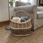 Oval wicker blanket basket holding folded knitted throws on a jute rug next to a linen armchair in a cosy living room