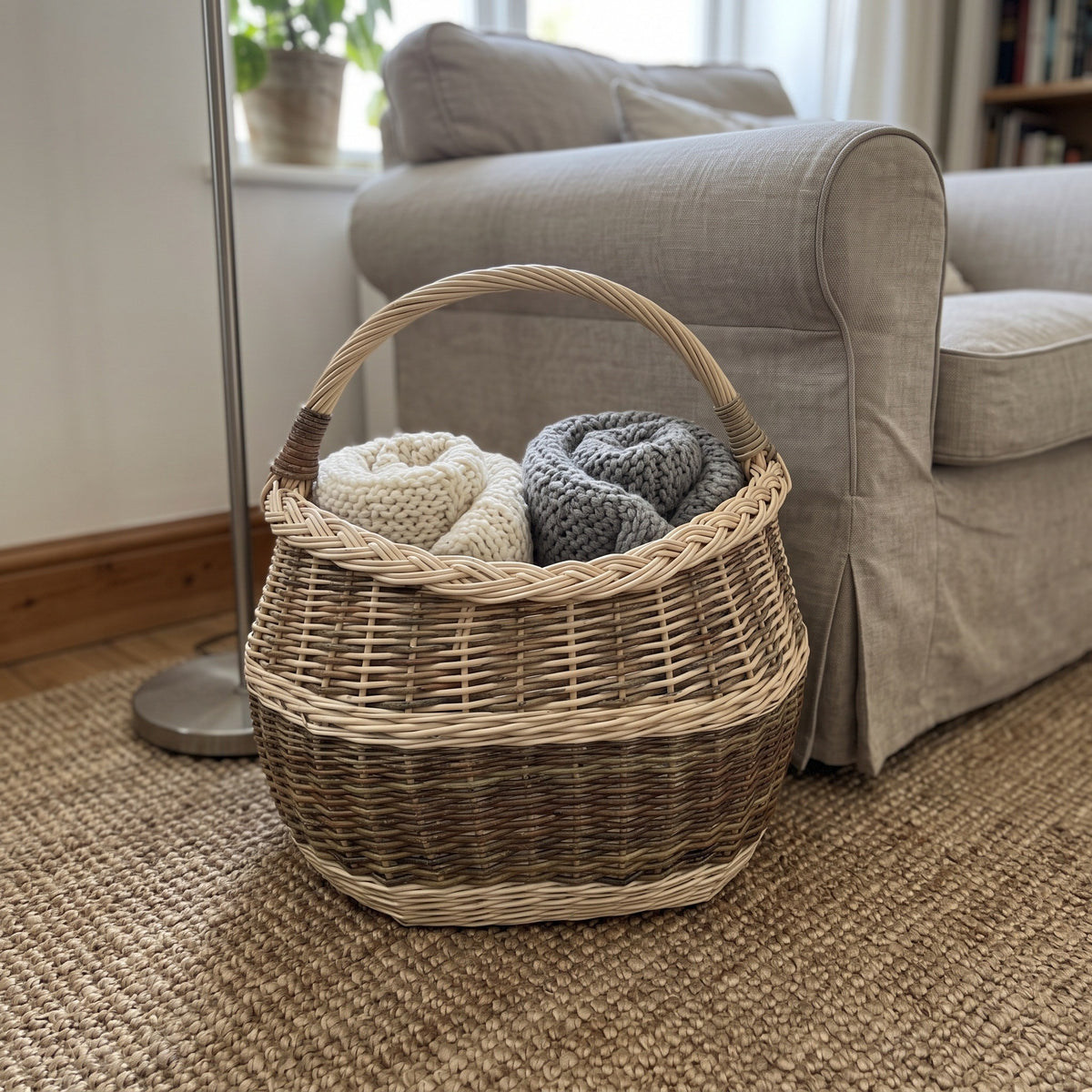 Oval wicker blanket basket holding folded knitted throws on a jute rug next to a linen armchair in a cosy living room