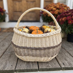 Oval wicker basket with a handle holding small pumpkins, gourds and pine cones on a wooden porch beside potted chrysanthemums