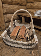 Handwoven willow log tray with an arched handle holding split firewood and kindling on a wooden floor inside a log cabin.