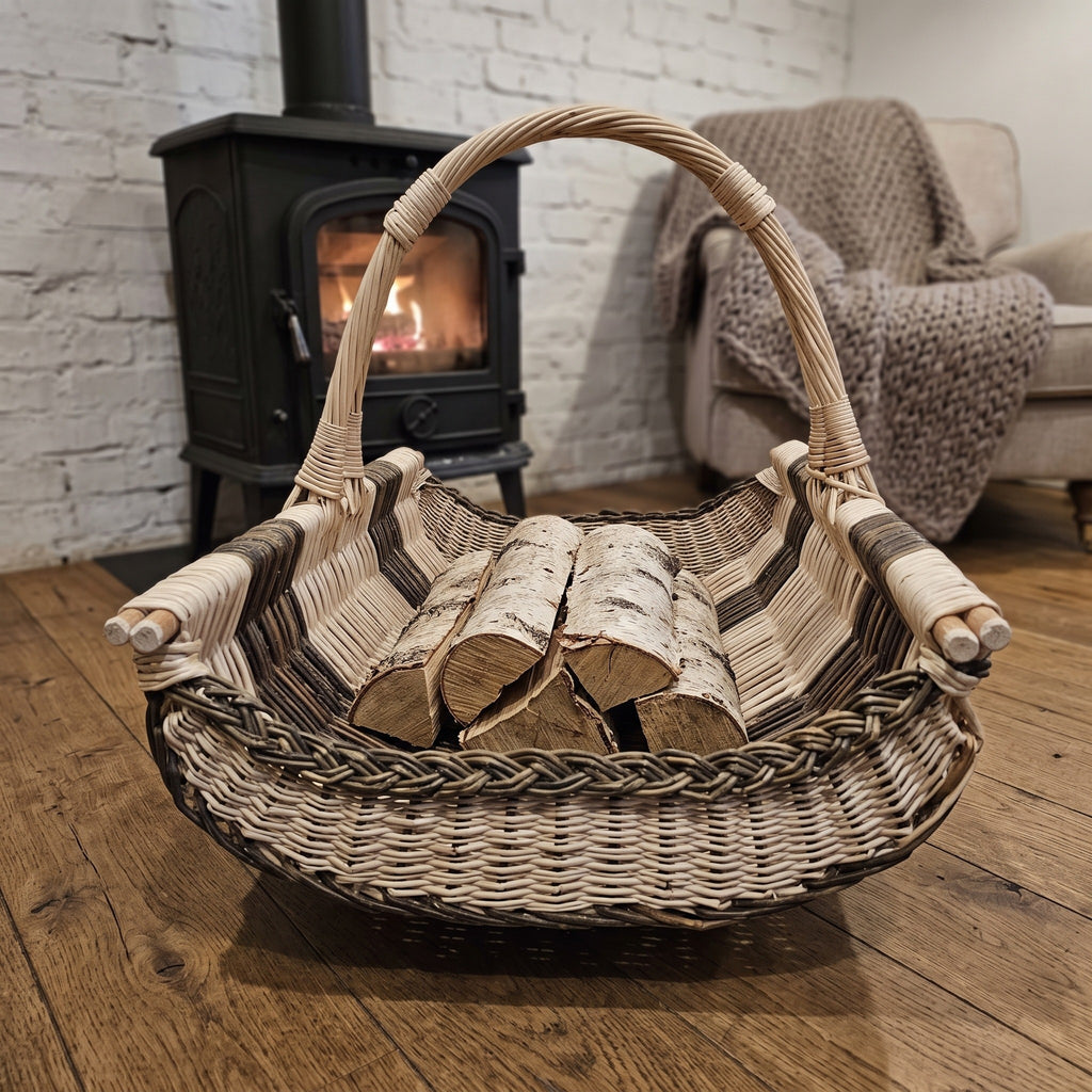 Willow log basket with a tall arched handle filled with birch logs on a wooden floor beside a wood stove and an armchair with a knitted throw.