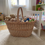 Large handwoven wicker basket with handle used for toy storage, filled with teddy bears and wooden blocks on a rug beside a child's bed