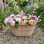 Handwoven wicker harvest basket with an arched handle overflowing with garden roses and peonies on a gravel path in a cottage flower garden