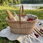 Oval wicker picnic carrier woven from willow with a baguette, cheese, cherries and a bottle of lemonade on a linen blanket by a lake