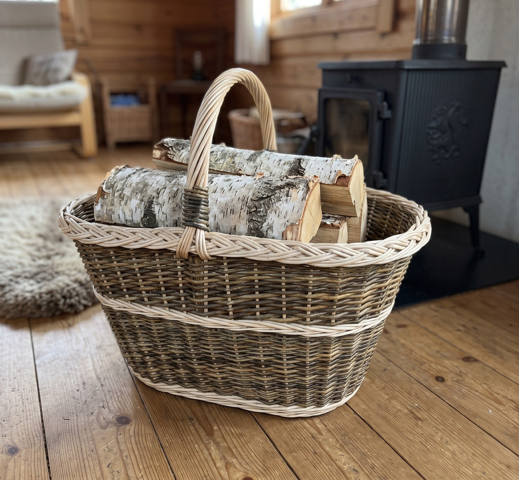 Large handwoven willow basket with handle used as a log basket, holding split birch logs on a plank floor beside a wood-burning stove
