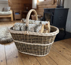 Large handwoven willow basket with handle used as a log basket, holding split birch logs on a plank floor beside a wood-burning stove