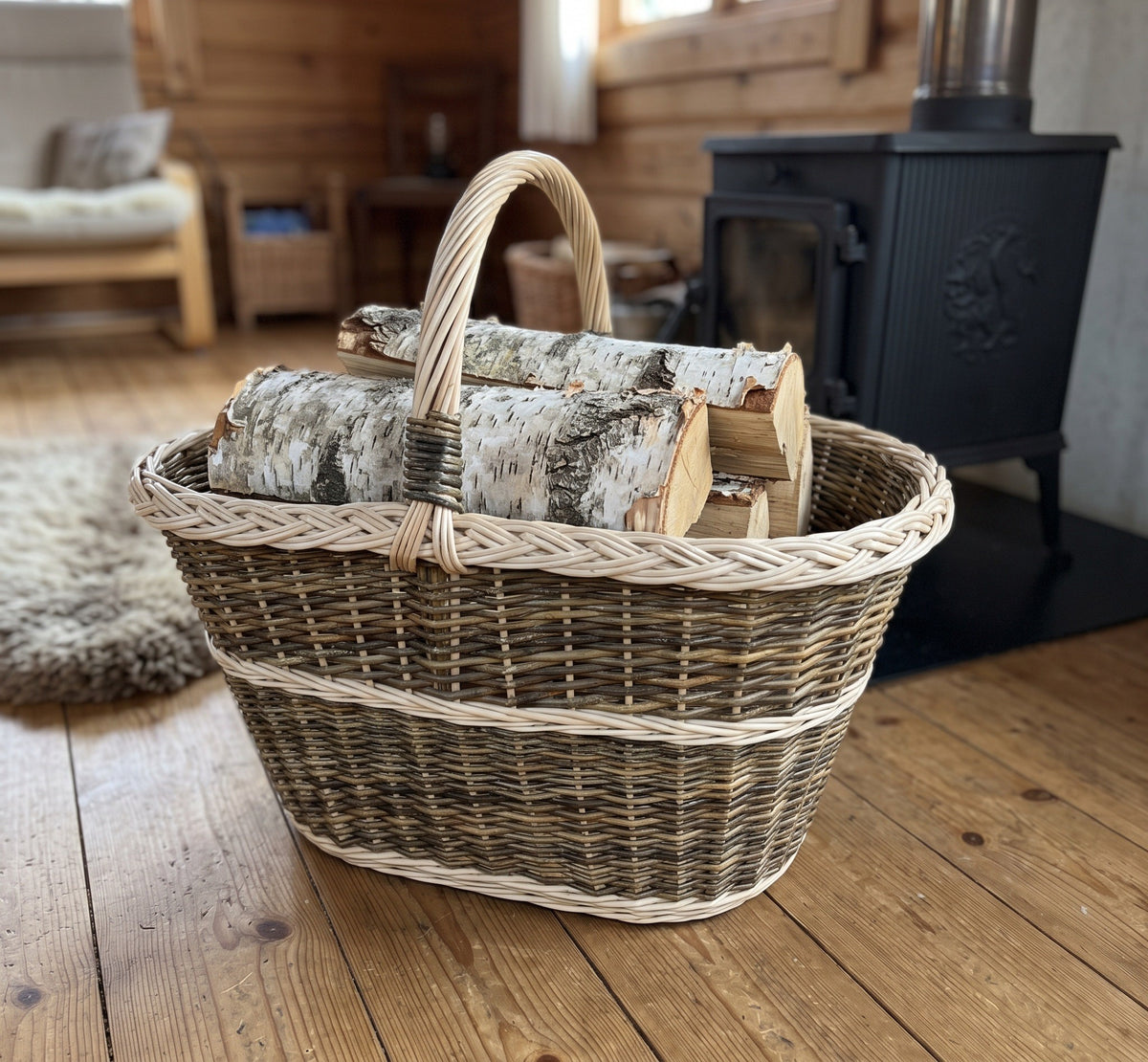 Large handwoven willow basket with handle used as a log basket, holding split birch logs on a plank floor beside a wood-burning stove