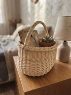 Small oval willow basket with handle holding books, glasses and a succulent on a wooden bedside table next to a lamp