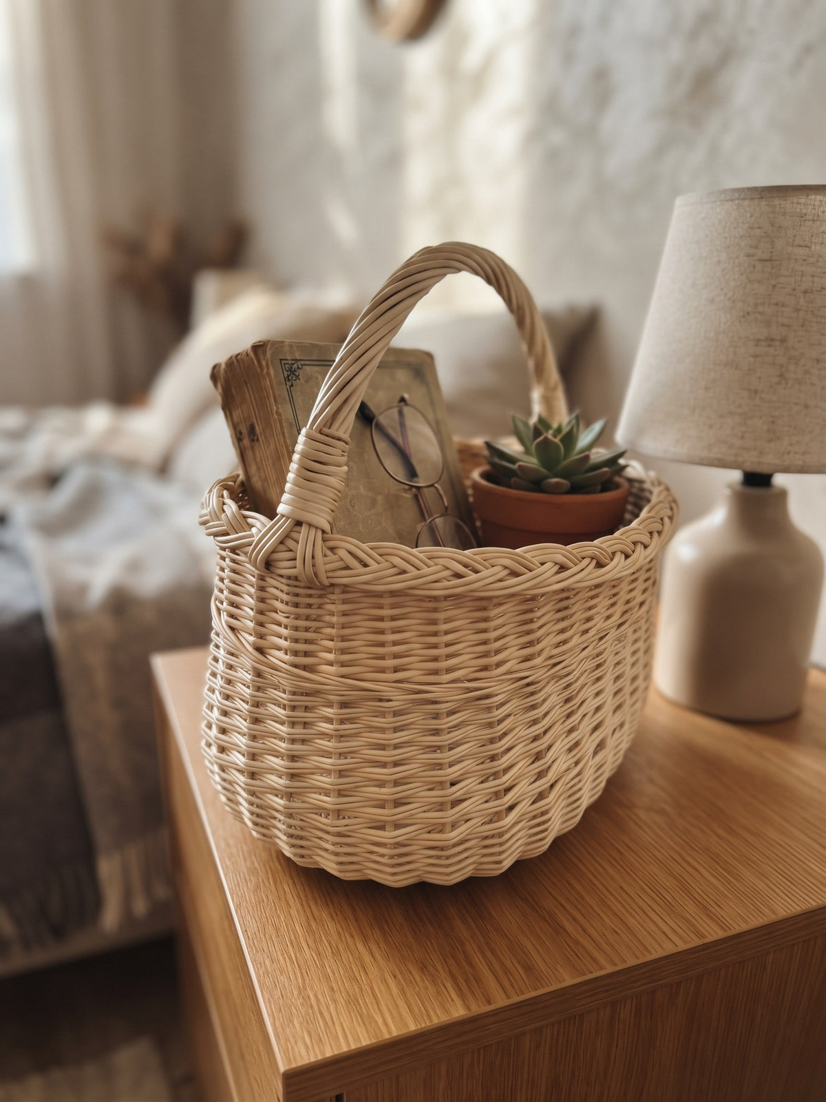 Small oval willow basket with handle holding books, glasses and a succulent on a wooden bedside table next to a lamp
