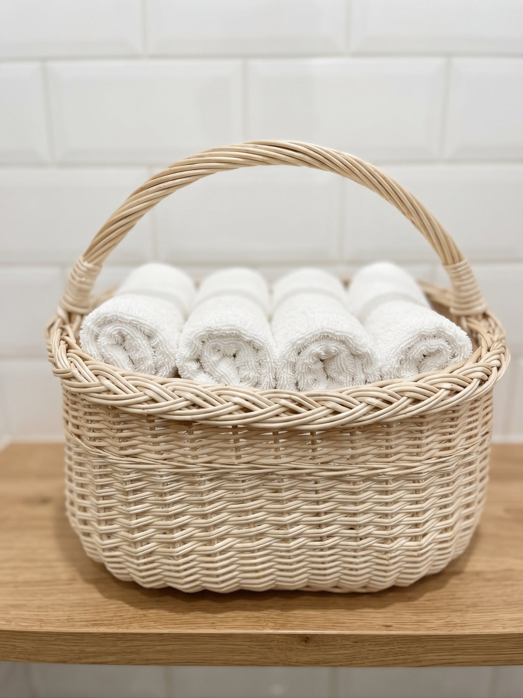 Small oval wicker basket with handle holding rolled hand towels on a wooden bathroom shelf against a tiled wall