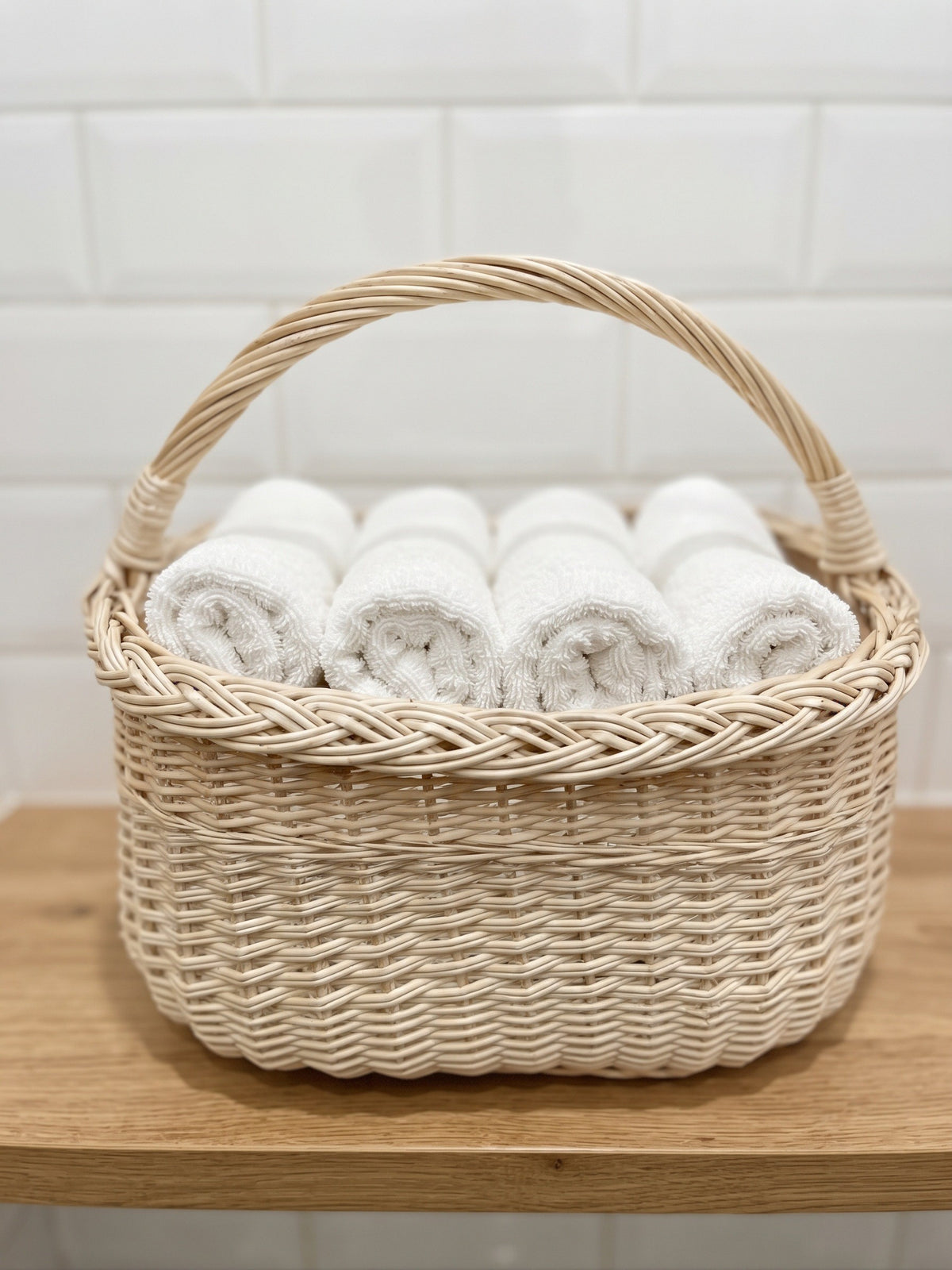 Small oval wicker basket with handle holding rolled hand towels on a wooden bathroom shelf against a tiled wall