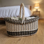 Oval wicker shopping basket with handle, handwoven from willow and filled with folded knitted blankets and linens on a wooden bedroom floor.