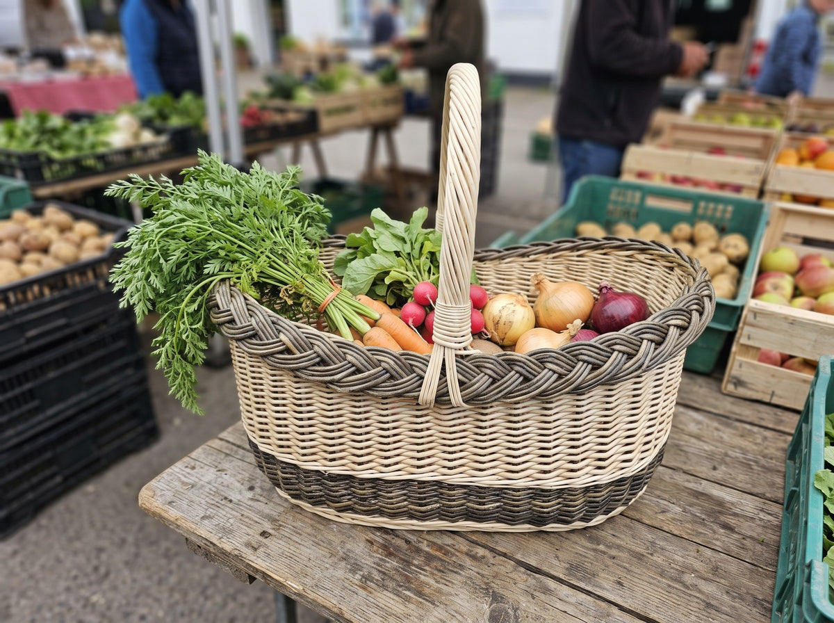 Handwoven willow market basket with arched handle holding carrots, radishes, onions and apples on a farmers market stall with produce crates.