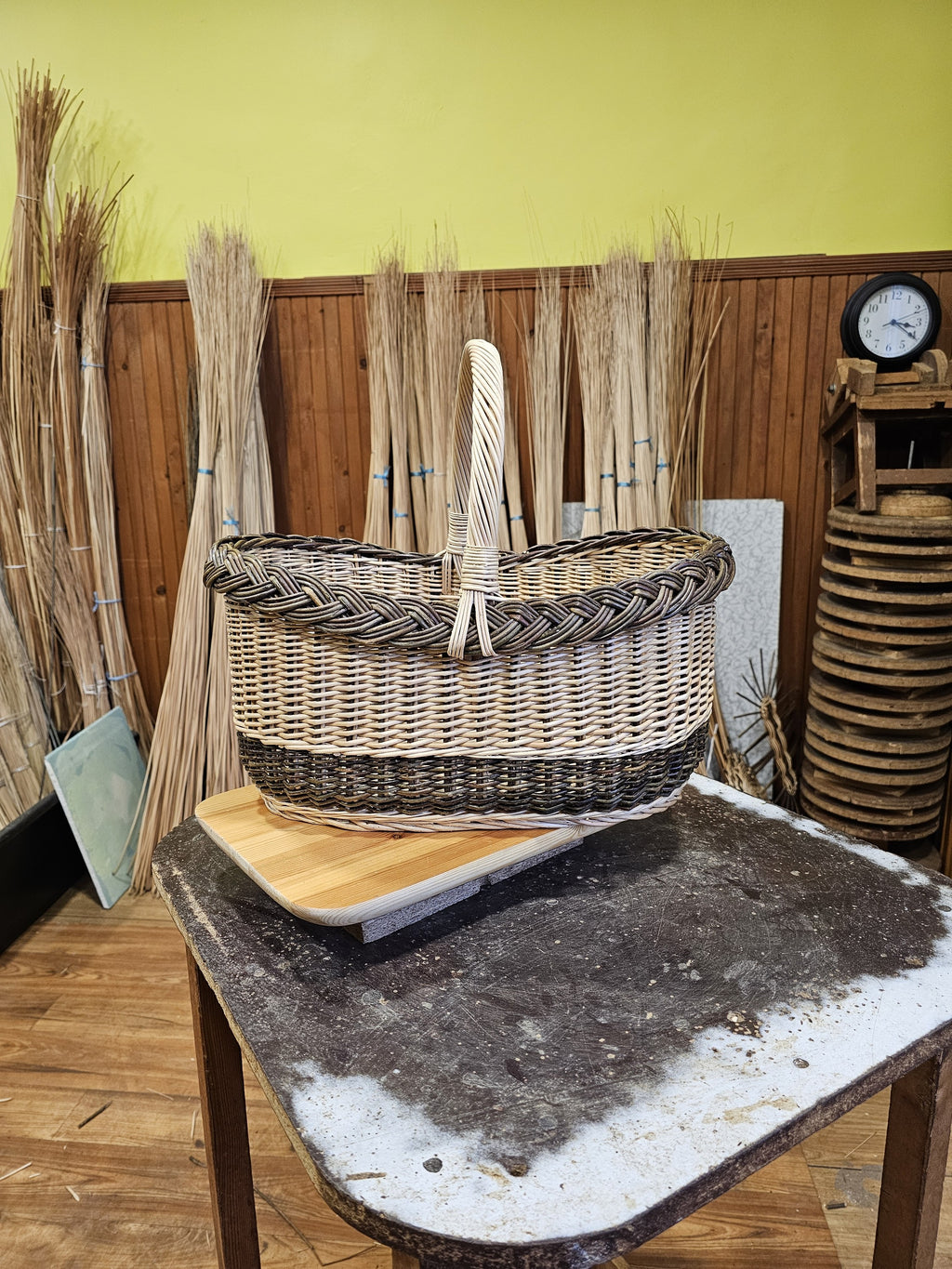 Oval willow shopping basket on a wooden cutting board in the workshop, with bundles of willow rods and a wall clock behind.