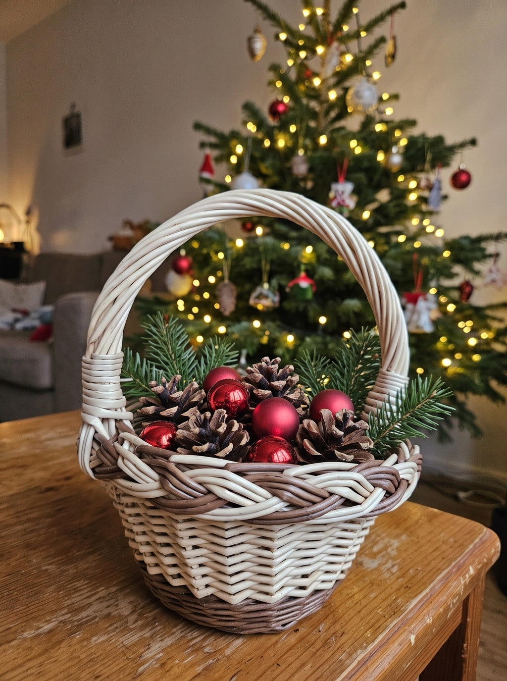 Mini handwoven willow basket with a braided rim filled with pine cones, red baubles and fir sprigs on a table before a lit Christmas tree.