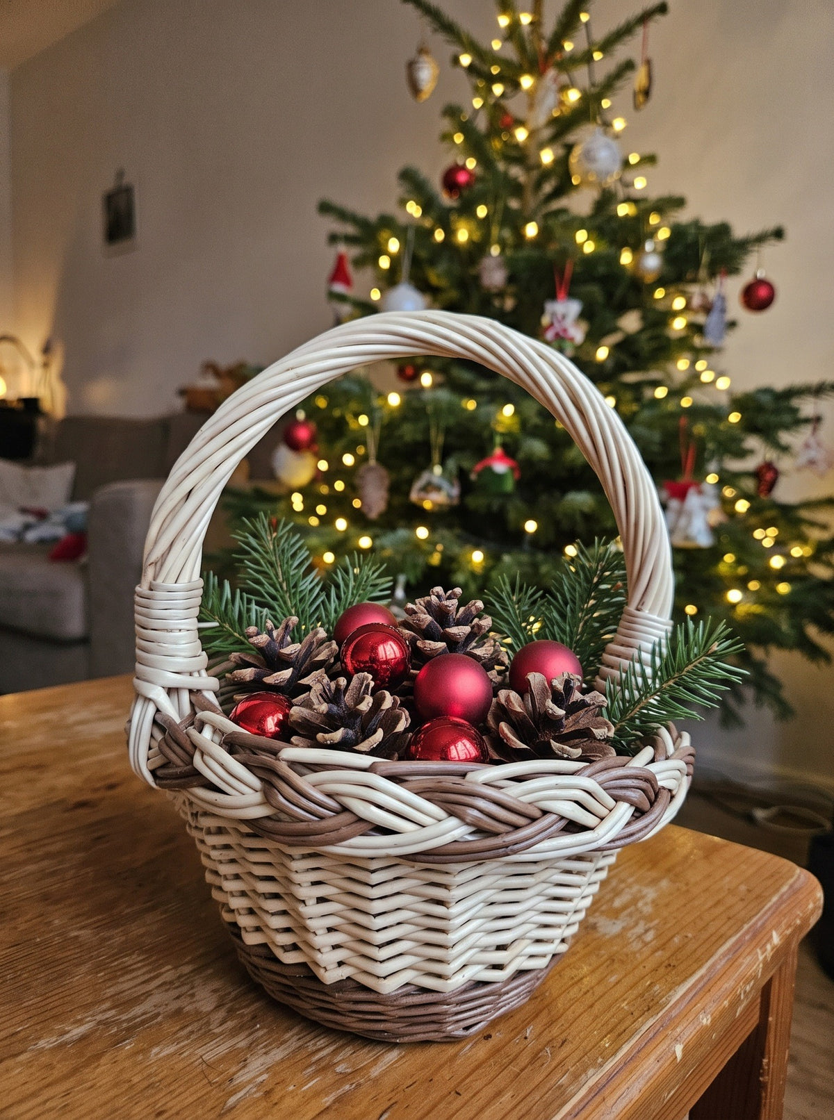 Mini handwoven willow basket with a braided rim filled with pine cones, red baubles and fir sprigs on a table before a lit Christmas tree.
