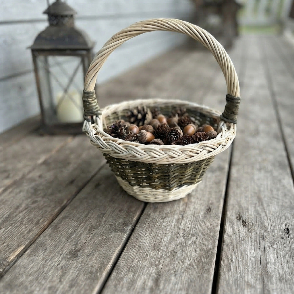 Small round wicker basket with a bound handle and braided rim holding pine cones on a weathered wooden porch beside a lantern.
