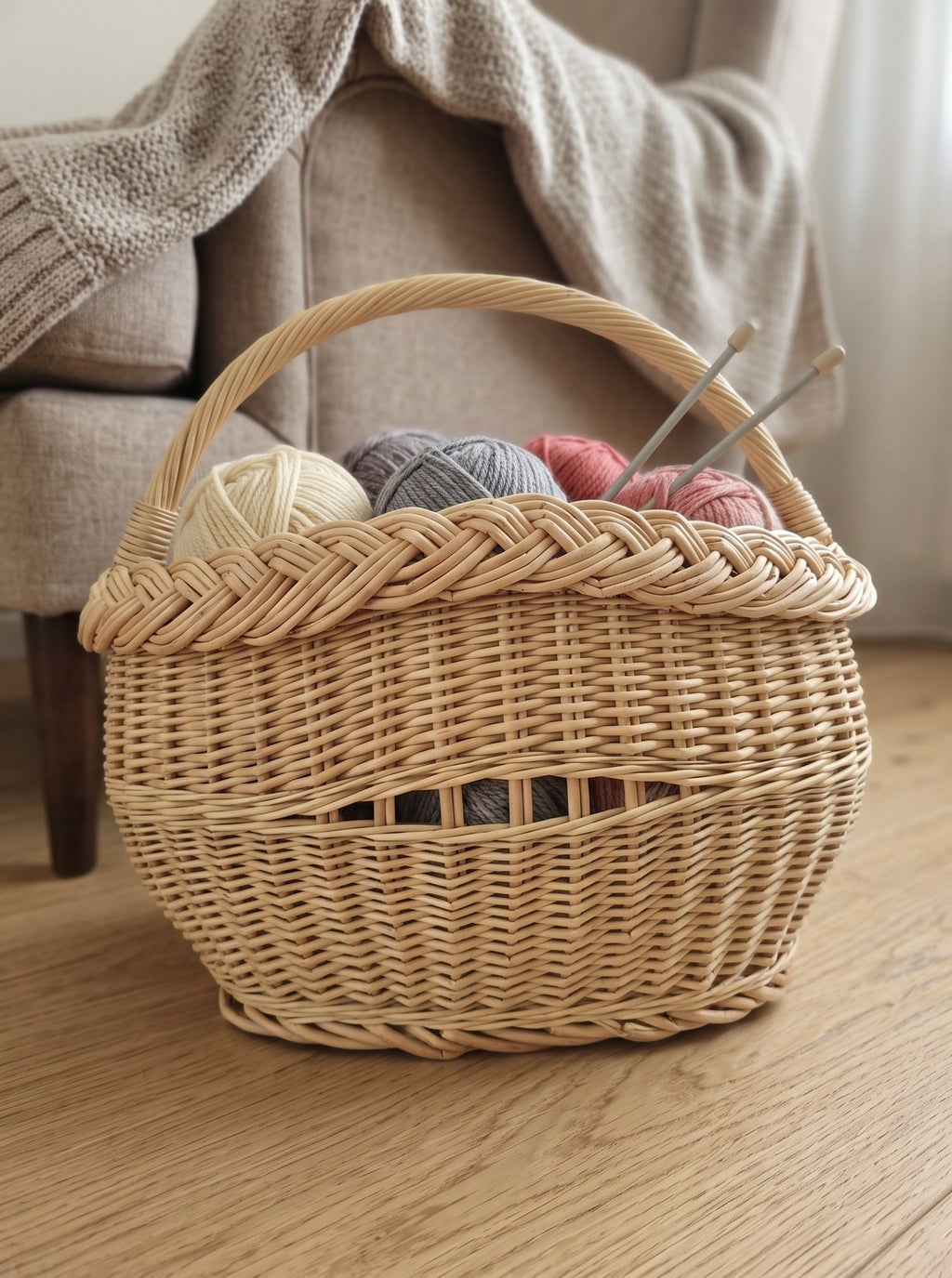 Small oval wicker knitting basket with handle holding balls of yarn and knitting needles on a wooden floor beside an armchair