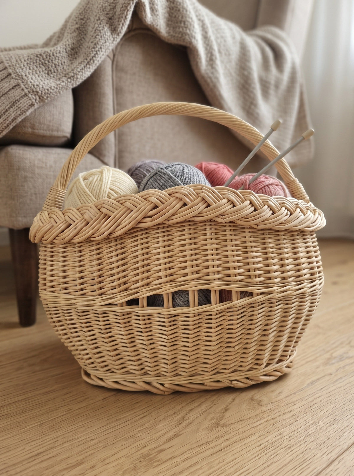 Small oval wicker knitting basket with handle holding balls of yarn and knitting needles on a wooden floor beside an armchair