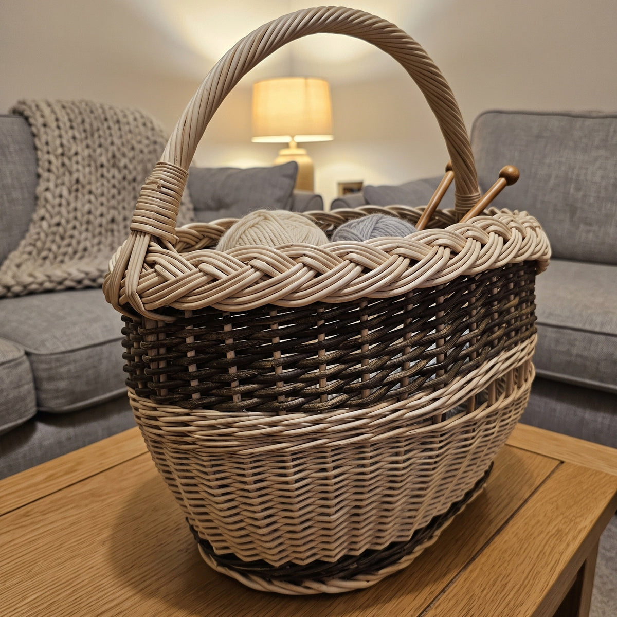 Oval wicker yarn basket with a braided rim holding balls of wool and knitting needles on a coffee table in a cosy living room