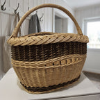 Small oval willow market basket with a braided rim and arched handle standing on a white shelf in a bright hallway
