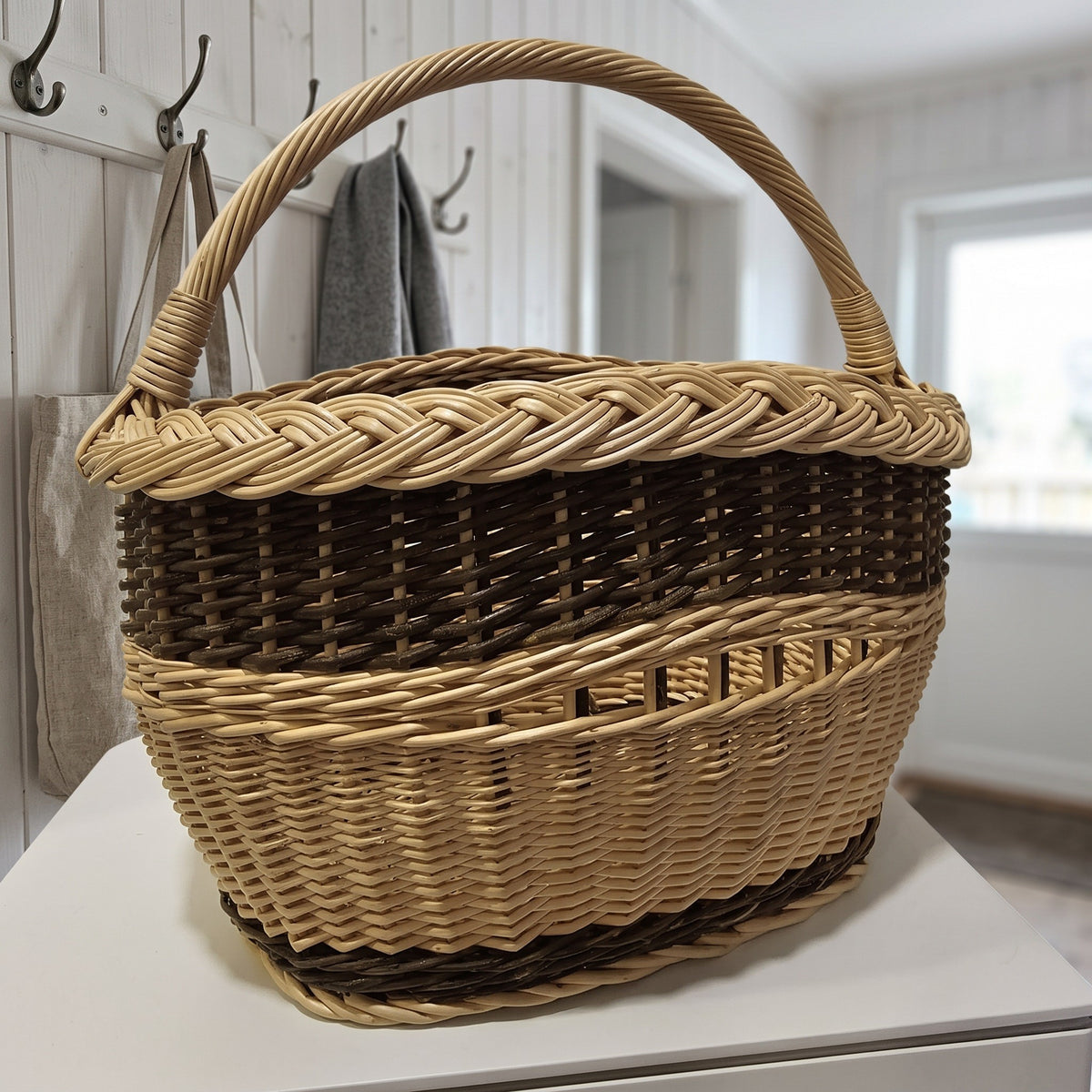 Small oval willow market basket with a braided rim and arched handle standing on a white shelf in a bright hallway