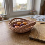 Small round handwoven willow bowl with a braided rim holding plums and apricots on a wooden kitchen table near the window