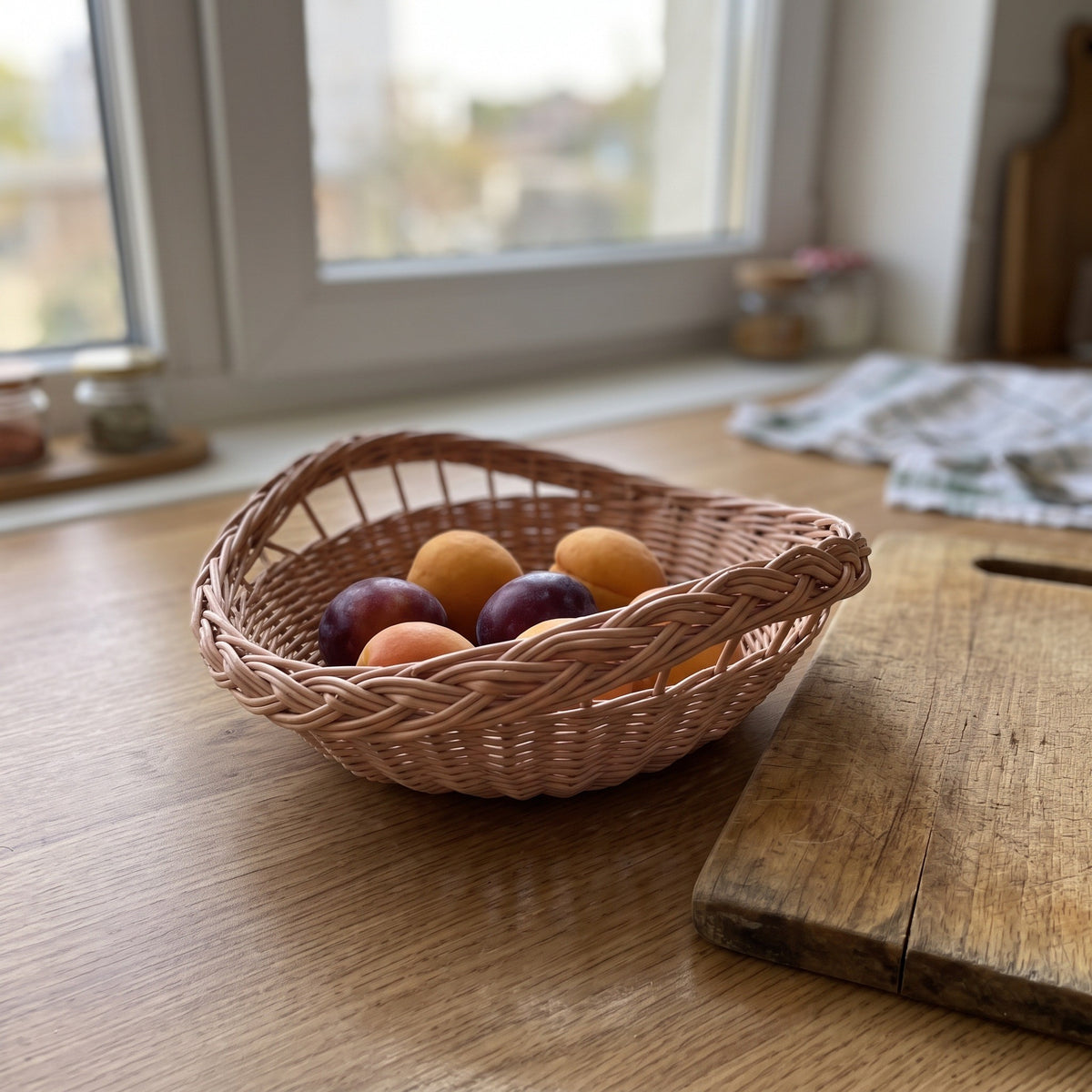Small round handwoven willow bowl with a braided rim holding plums and apricots on a wooden kitchen table near the window