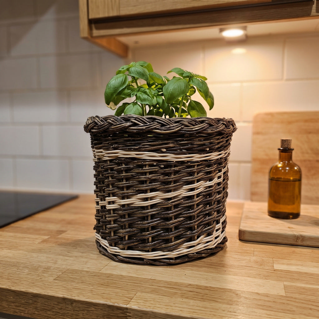 Indoor planter basket in the small size with a braided rim holding a potted basil plant on a kitchen worktop beside a glass bottle.