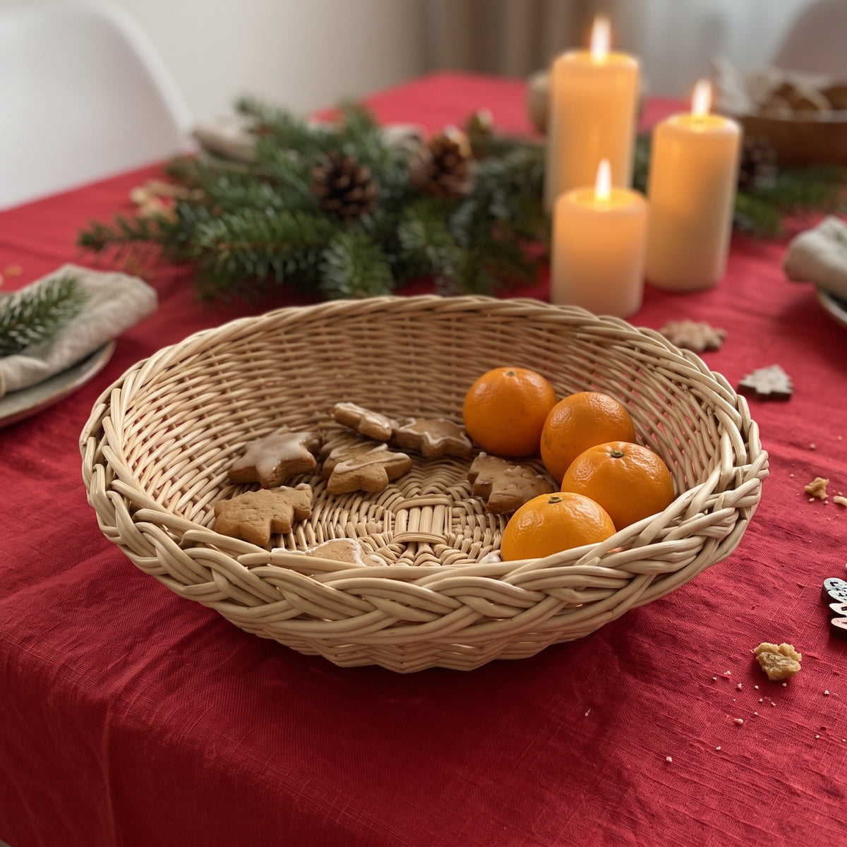Round wicker bread basket with a braided rim on a festive Christmas table, holding gingerbread cookies and tangerines beside lit candles and pine branches.