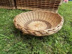 Handwoven willow bread basket resting on grass beside larger wicker baskets, showing its braided rim and radial woven base.
