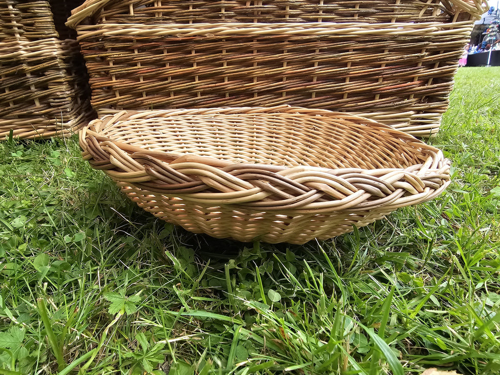 Side view of the shallow willow serving tray on grass, its thick braided rim and low profile shown against larger wicker baskets behind.