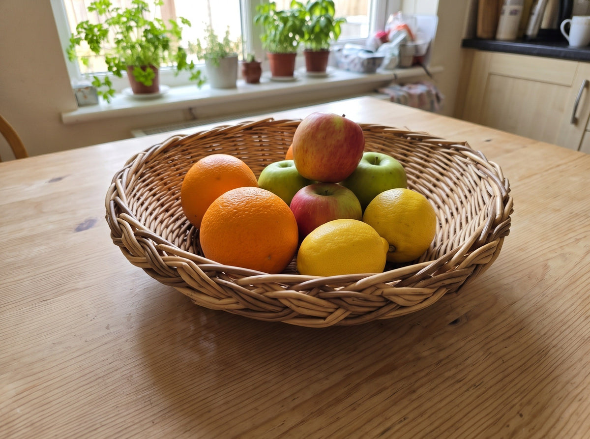 Handwoven willow fruit bowl with braided rim on a kitchen table by the window, filled with oranges, apples and lemons.