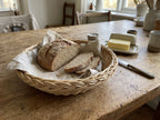 Round willow bread basket lined with a linen napkin, holding a sliced rye loaf on a farmhouse table with a butter dish and knife.